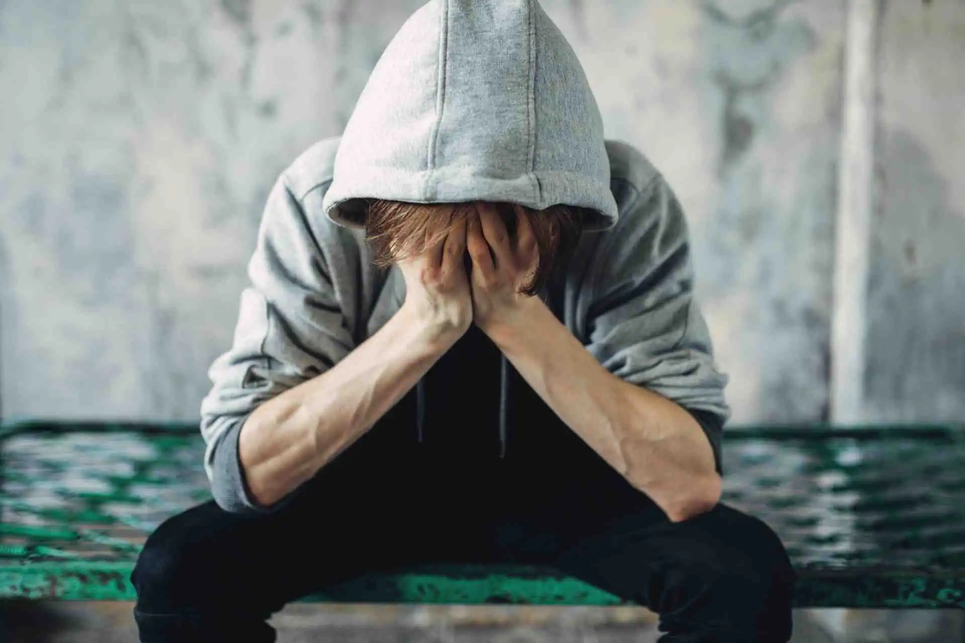 Person in a hoodie sitting with head in hands, showing emotional distress linked to early drug addiction